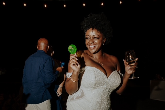 A woman in a white dress is smiling and holding a green lollipop in one hand and a glass of wine in the other. People are visible in the background under dim lighting.