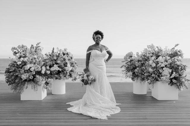 A woman in a wedding dress stands on a wooden deck, holding a bouquet, with large floral arrangements and the ocean in the background. (black and white image).