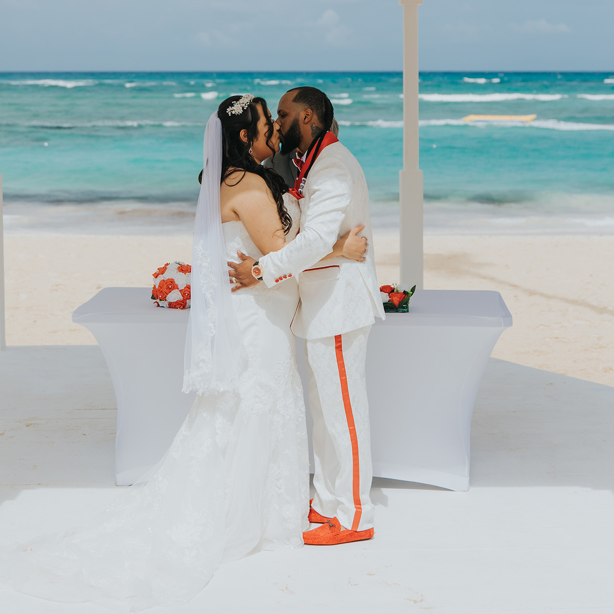 A couple dressed in wedding attire shares a kiss in front of an outdoor setup with a white table and the ocean in the background.