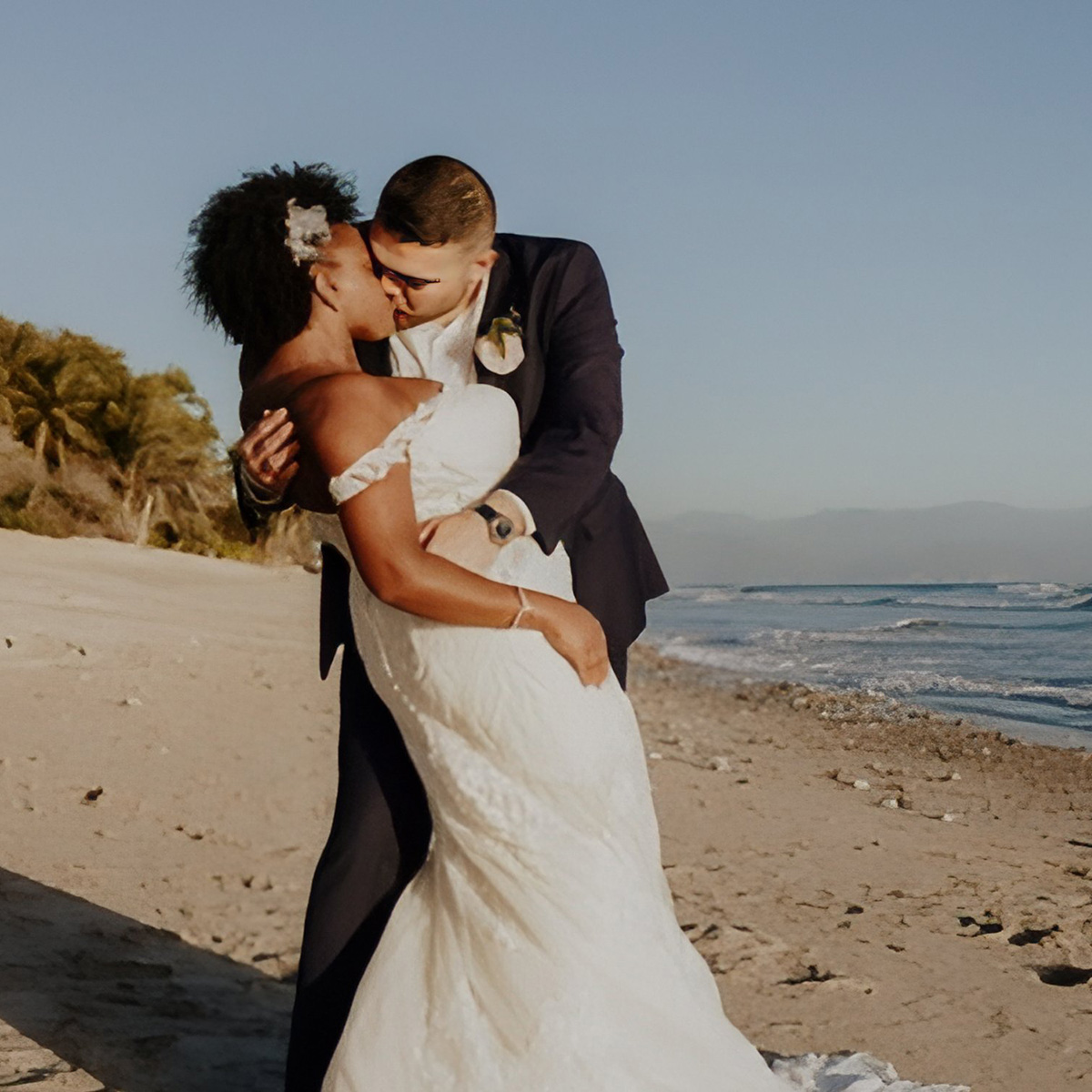 A couple in a wedding dress and suit share a kiss on a beach with the sea and palm trees in the background.