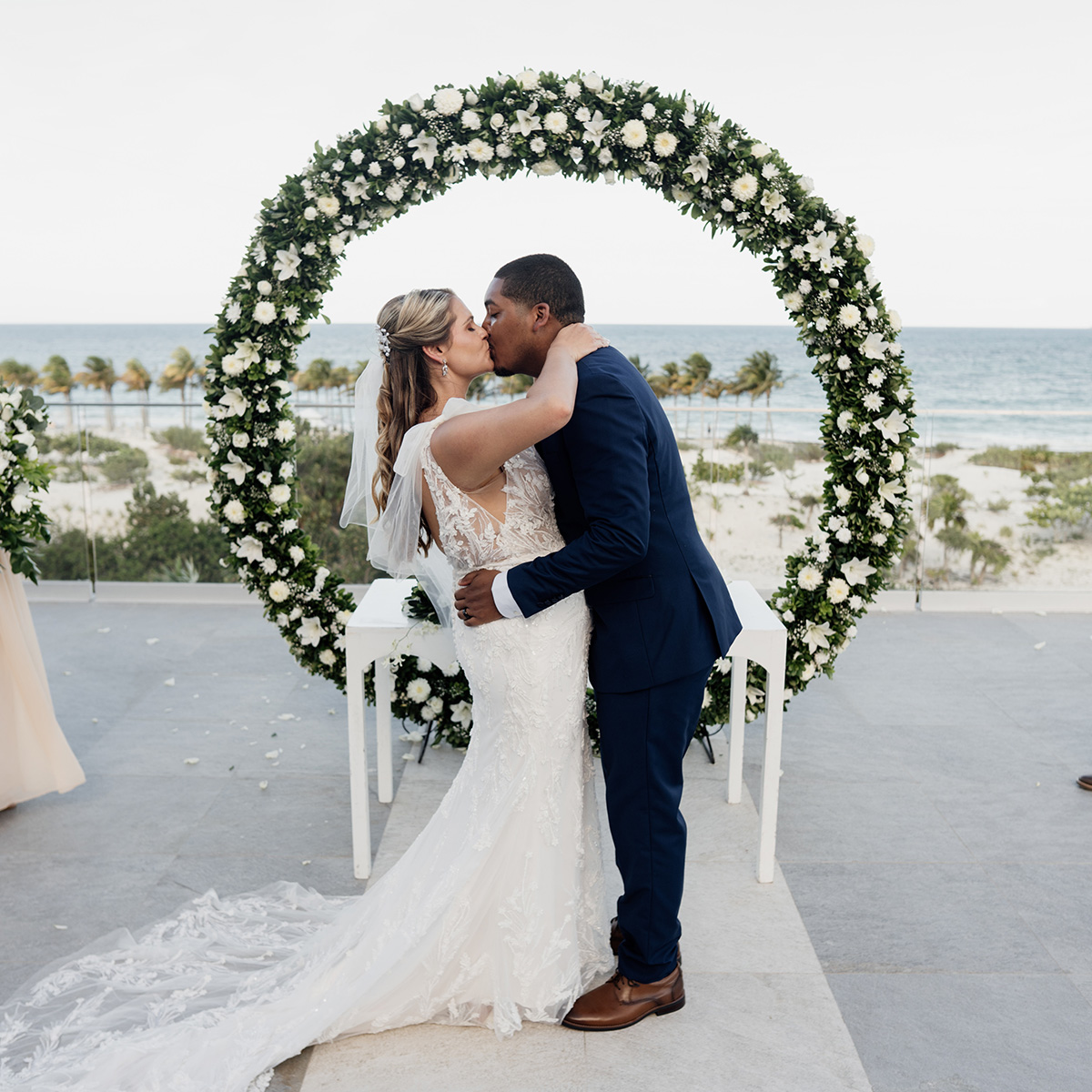 Lauren and Julian share a kiss under a grand floral arch by the ocean, with the bride in a white gown and the groom wearing a navy suit.