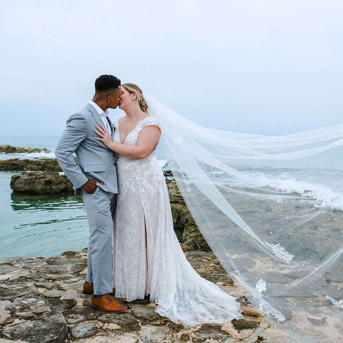 Mollie Schillinger marries her groom in a light gray suit, sharing a kiss on rocky terrain near the ocean.