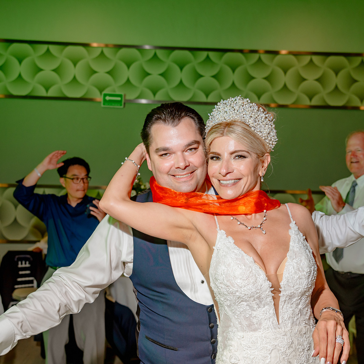 A man and a woman dressed in formal attire are smiling and posing together. The woman, wearing the "Nancy" white lace dress adorned with a crown, stands beside the man who is sporting a vest and shirt complemented by an orange scarf around his neck.