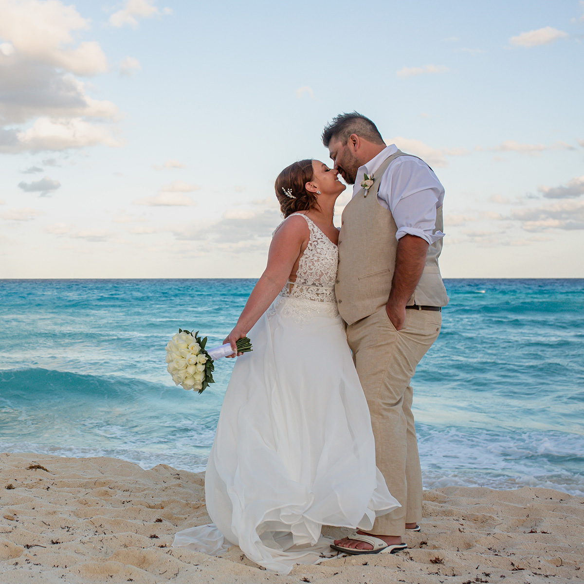 A bride and groom share a kiss on a sandy beach by the ocean, with the bride holding a bouquet of white flowers.