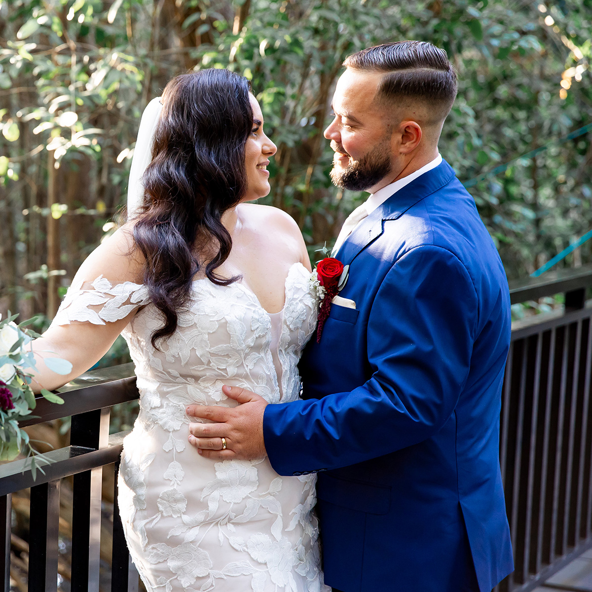 Annellis Anduja in wedding attire stand close together on an outdoor balcony, smiling at each other. Annellis wears a white dress with lace details, and Anduja is dressed in a blue suit with a red rose boutonniere.