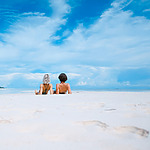 Two people in swimsuits lie on a white sandy beach, facing the ocean under a partly cloudy blue sky.