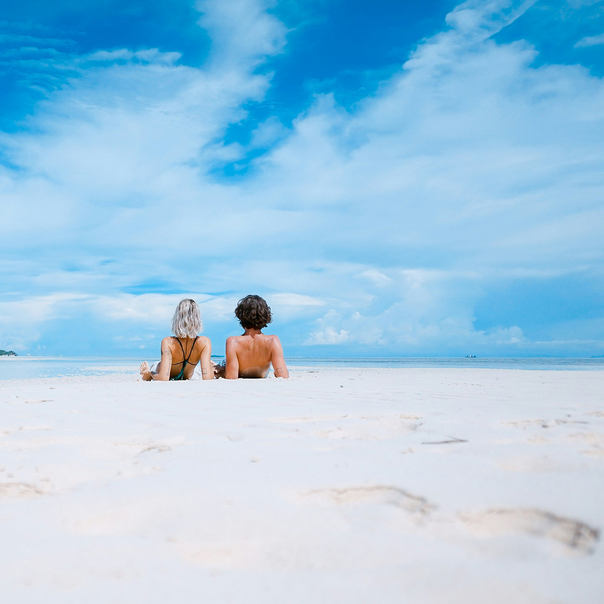 Two people in swimsuits lie on a white sandy beach, facing the ocean under a partly cloudy blue sky.