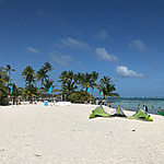 A sunny beach with white sand, palm trees, and clear blue skies. People are relaxing and walking near the shoreline, while large tents are set up on the sand. The ocean is visible in the background.