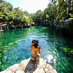 A person in a patterned swimsuit sits on a rock by a clear green natural water body surrounded by lush greenery, under a bright sunny sky.