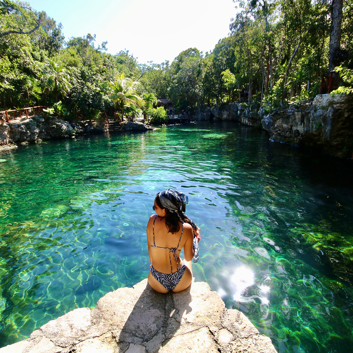 A person in a patterned swimsuit sits on a rock by a clear green natural water body surrounded by lush greenery, under a bright sunny sky.