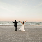 A bride and groom stand on a beach facing the ocean with their arms raised in celebration.