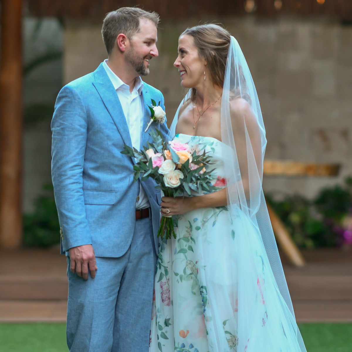 Jenna, wearing a floral wedding dress and veil, stands holding a bouquet, gazing at Ray in his light blue suit outdoors.