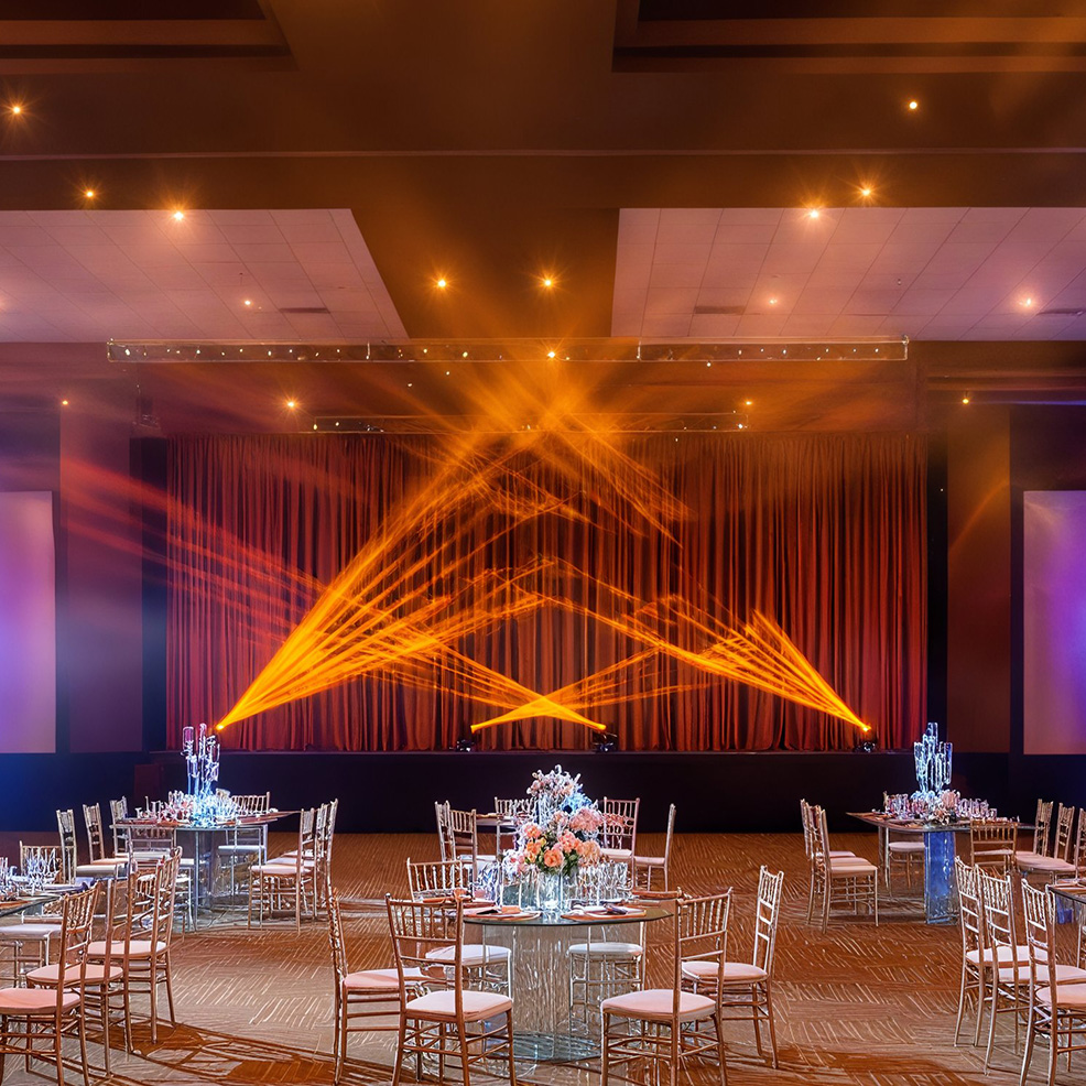 Ballroom with round tables and chairs arranged, floral centerpieces, and a stage with orange lighting and draped curtains.