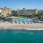 Beachfront resort with multiple buildings, a large swimming pool, lounge chairs under umbrellas, palm trees, and the ocean in the foreground.