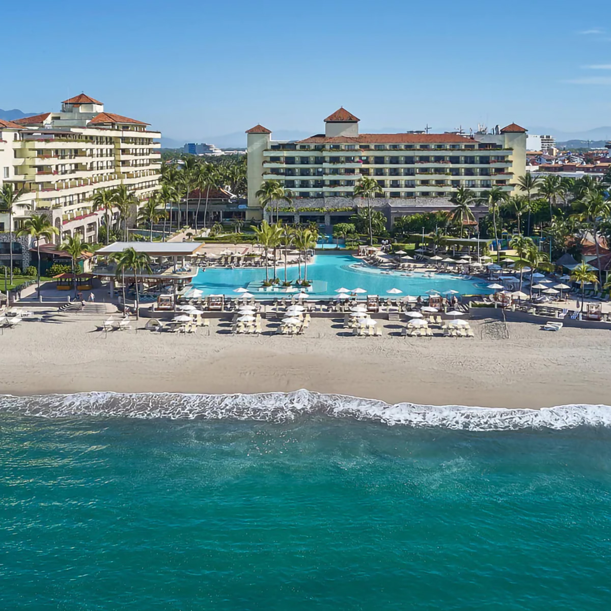 Beachfront resort with multiple buildings, a large swimming pool, lounge chairs under umbrellas, palm trees, and the ocean in the foreground.