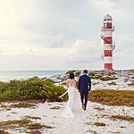 A couple in formal attire walks hand-in-hand toward a red and white lighthouse on a sandy beach, perhaps envisioning their dream celebration with one of the Mexico wedding packages for 75 guests.