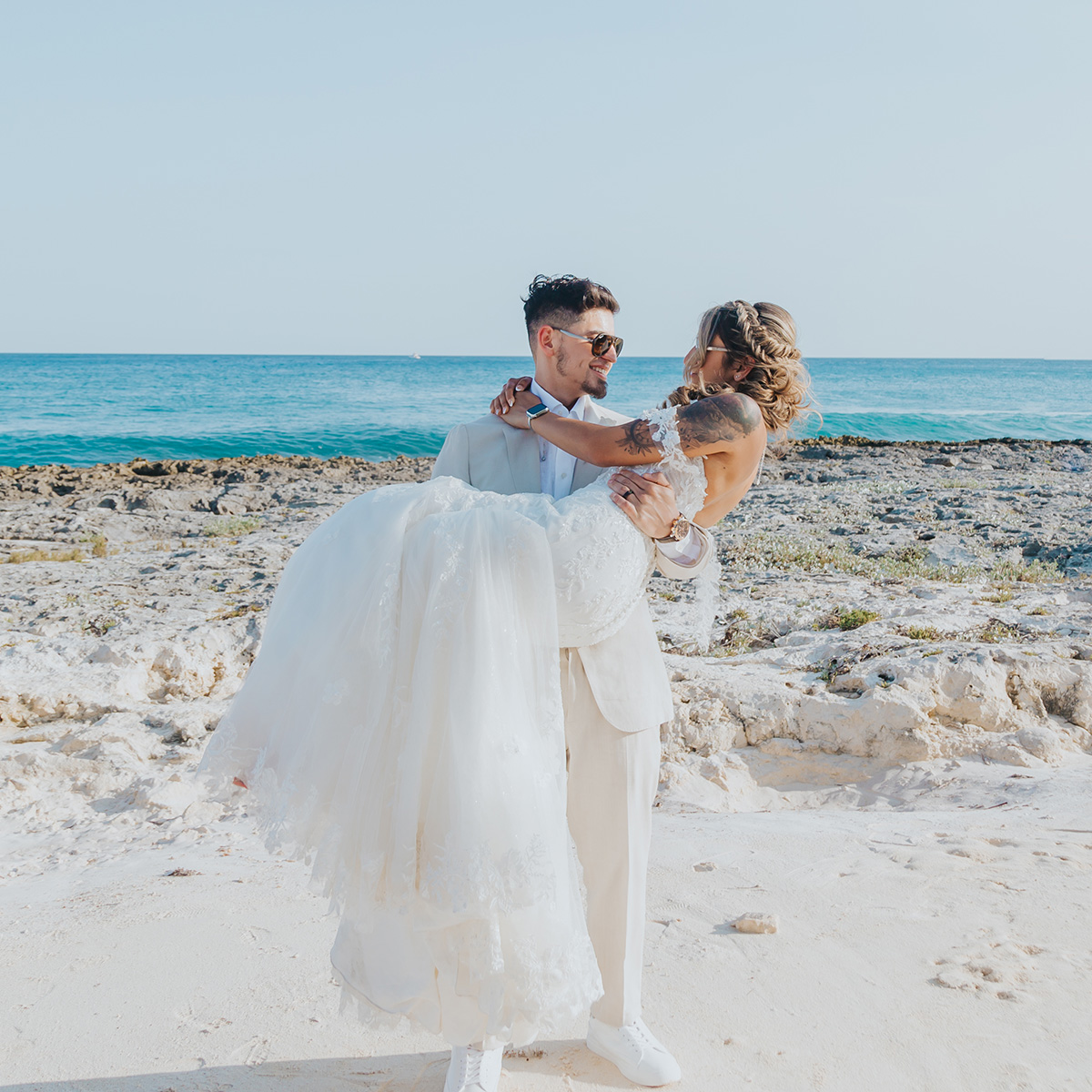 A groom in a cream suit, named Christian, lifts his bride, Nadia in a white wedding dress on a rocky beach with the ocean in the background.