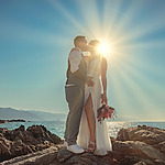 A couple kisses while standing on rocks by the ocean during sunset, with the sun positioned directly behind them, creating a backlit silhouette effect. The woman holds a bouquet of flowers.