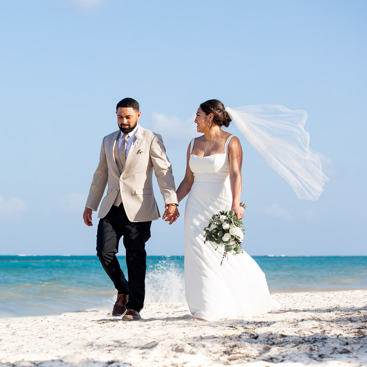 Samantha Bejarano and her groom hold hands as they walk along a sandy beach, with the ocean and a clear sky in the background. Samantha carries a bouquet, and her veil flows gracefully in the wind.