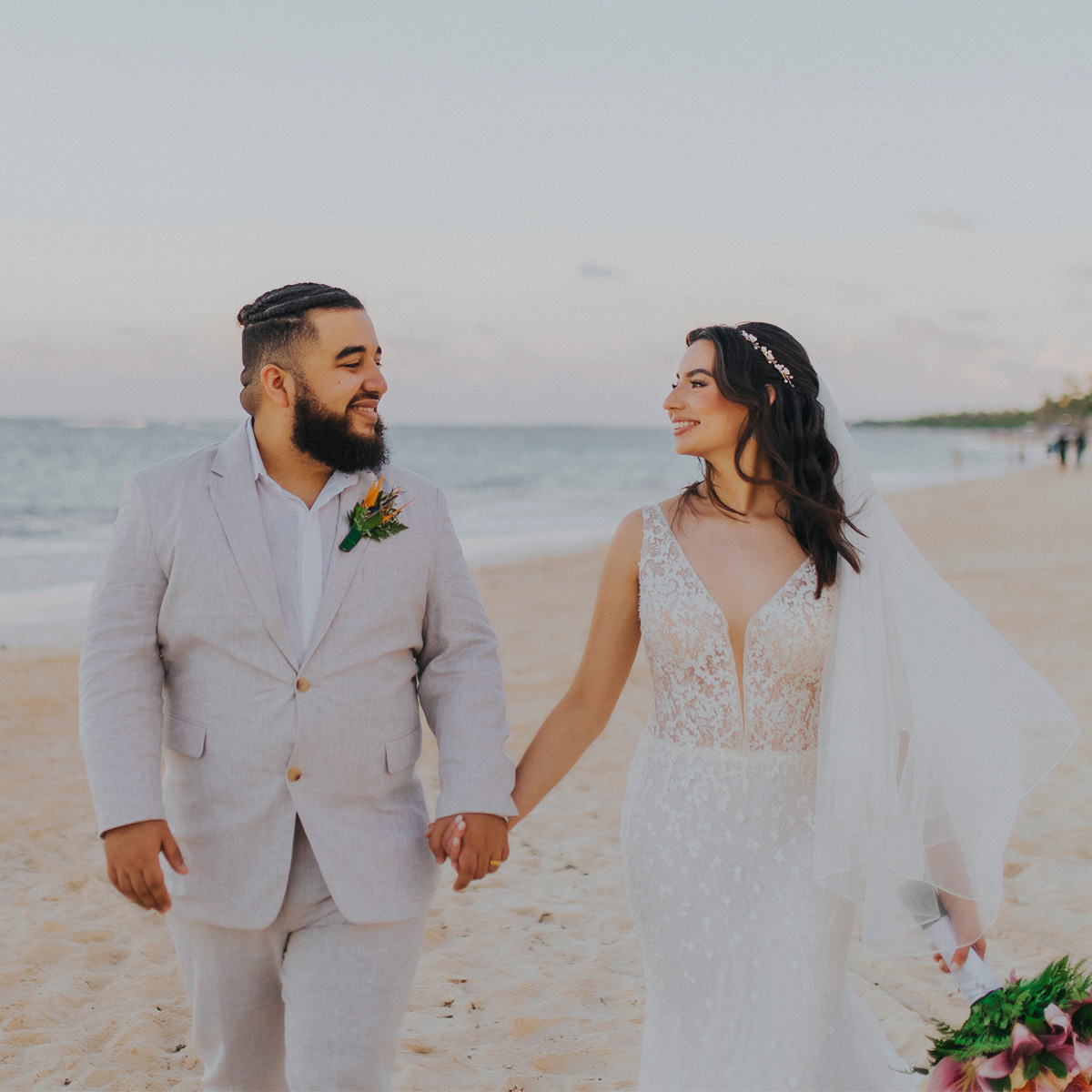 Solange and Joshua walk hand in hand on a sandy beach. Joshua is in a light gray suit, while Solange wears a white lace dress with a veil. The ocean and sky create a stunning backdrop.