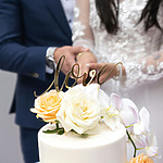 A bride and groom, dressed in white and blue, are cutting a wedding cake adorned with white and peach roses and a gold "love" topper.