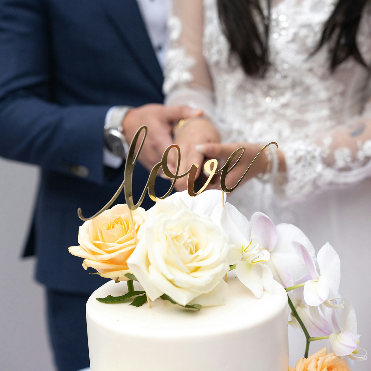 A bride and groom, dressed in white and blue, are cutting a wedding cake adorned with white and peach roses and a gold "love" topper.