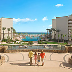 A family of four walks toward a large resort pool area, surrounded by palm trees, with a view of the ocean in the background.