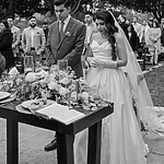A bride and groom stand at a decorated table with an open book during an outdoor wedding ceremony, surrounded by guests.