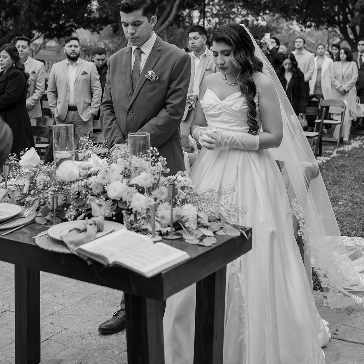 A bride and groom stand at a decorated table with an open book during an outdoor wedding ceremony, surrounded by guests.
