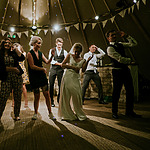 A group of people are dancing inside a tent decorated with string lights and bunting. The atmosphere is joyous and lively, with everyone engaged in the moment.