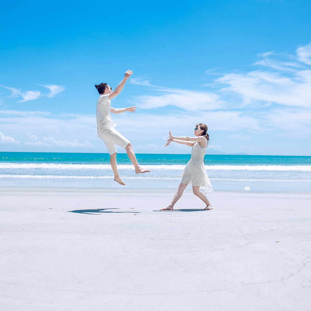 Two people in white outfits enjoy the beach; one person jumps in the air while the other reaches out with open arms under a clear blue sky.