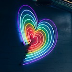 Neon artwork of a heart shape in a rainbow gradient, mounted on a dark wall with industrial piping above.
