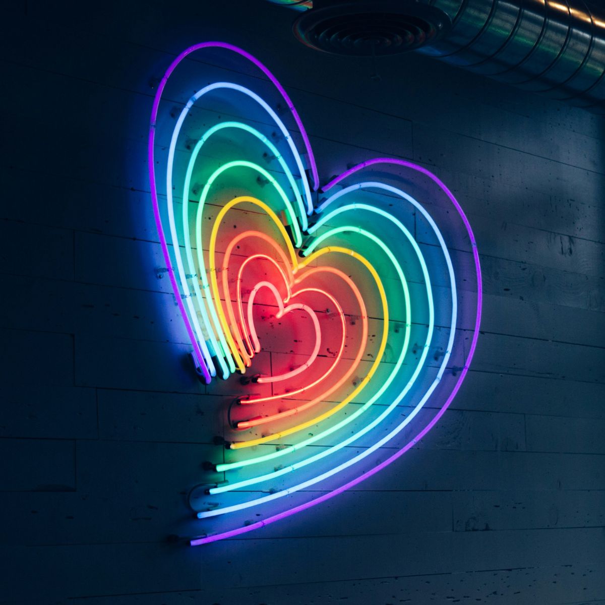 Neon artwork of a heart shape in a rainbow gradient, mounted on a dark wall with industrial piping above.