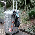 A silver suitcase adorned with colorful stickers stands on a paved path by greenery, with a black and blue umbrella attached to its handle.