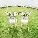 Two chairs adorned with flowers face a grassy area by water. Each chair has a sign; one reads "Forever" and the other "Always.