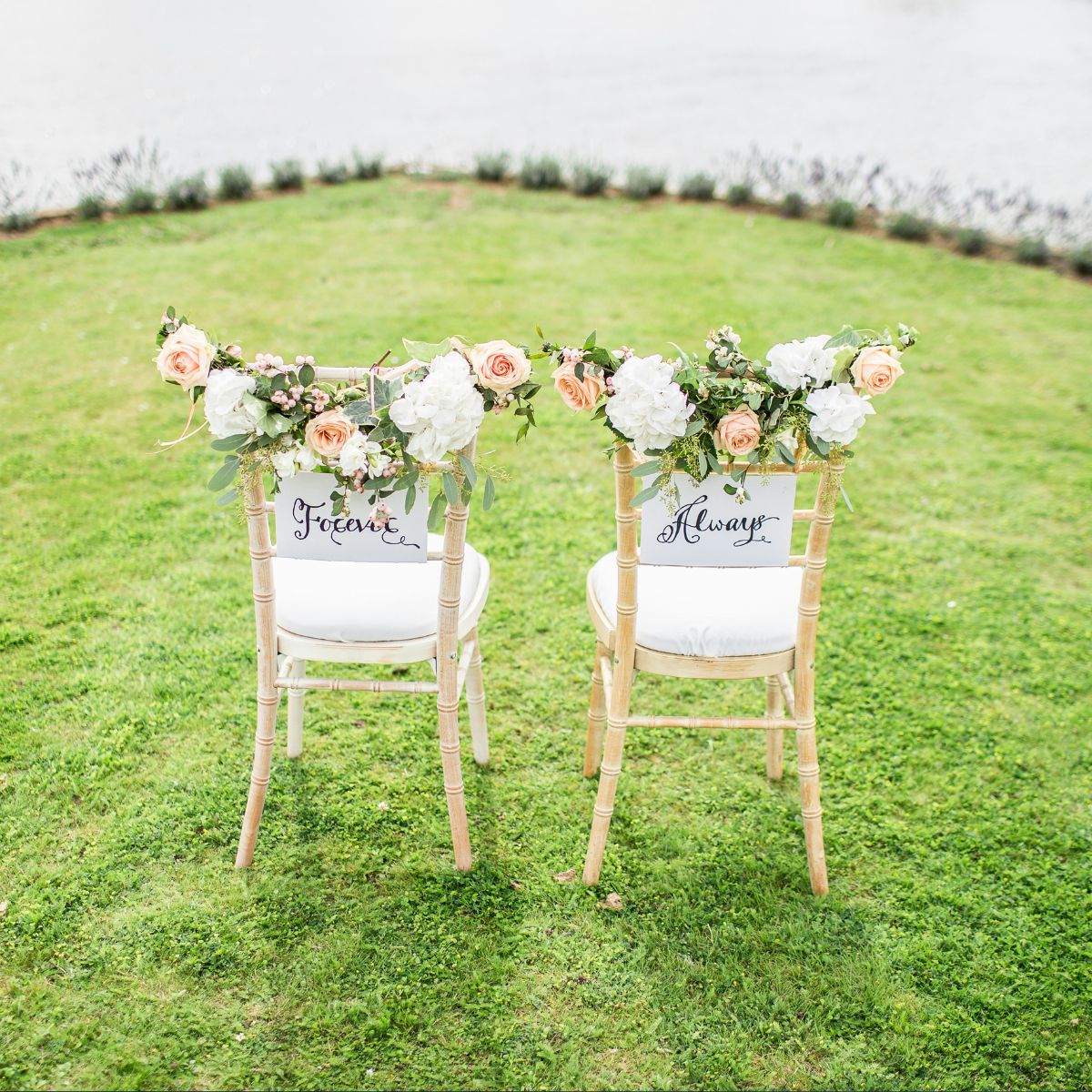 Two chairs adorned with flowers face a grassy area by water. Each chair has a sign; one reads "Forever" and the other "Always.