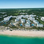 Aerial view of the Riu Palace Bavaro, a large beachside resort with multiple white buildings, several swimming pools, surrounded by lush green vegetation, and a long, sandy beach meeting turquoise water.