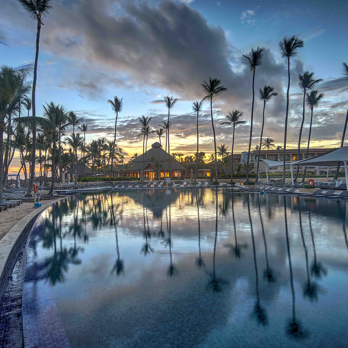 A large, calm swimming pool reflecting tall palm trees and a building at sunset, with lounge chairs arranged around the pool's edge.