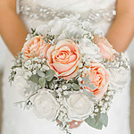 A bride holds a bouquet of peach and white roses accented with greenery and small white filler flowers.