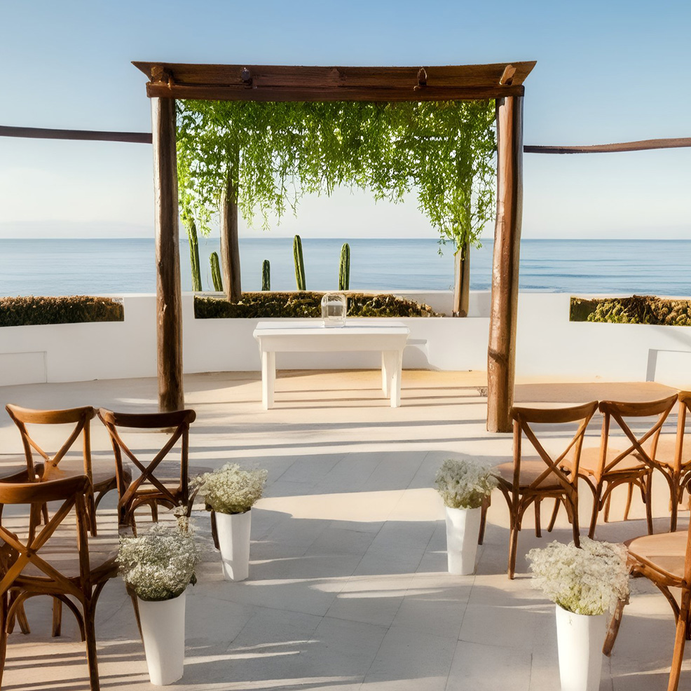 Outdoor wedding ceremony setup with wooden chairs, white flowers, a wooden arch, and a view of the ocean in the background.