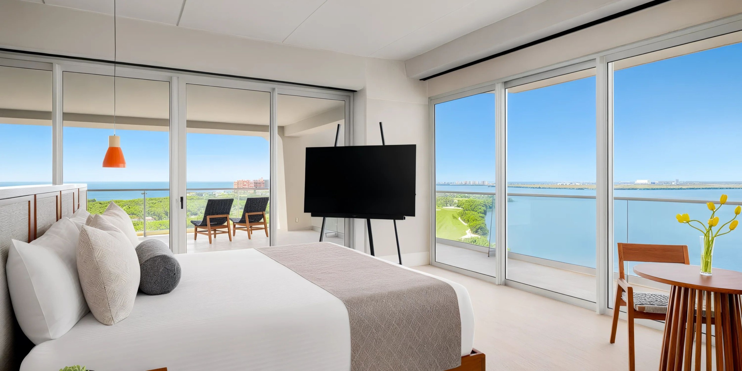The Hyatt Vivid Grand Island presents a contemporary hotel room featuring expansive windows that reveal a balcony with stunning ocean views. Inside, you'll find a comfortable bed, a TV set on a stand, a round table paired with chairs, and an elegant pendant light.