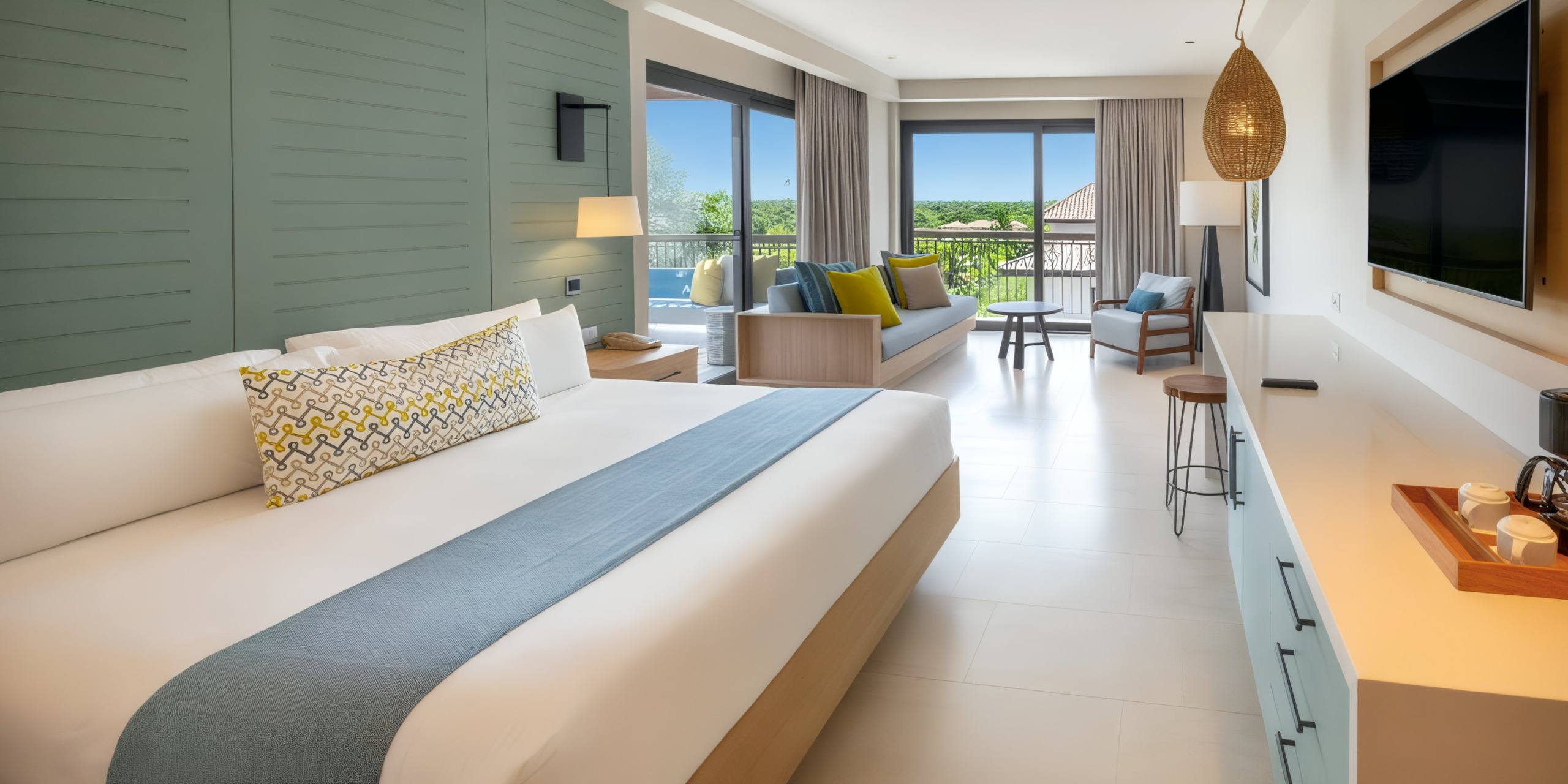 The Lopesan Costa Bavaro offers a spacious hotel room featuring a large bed, sofa, TV, and an outdoor view through glass doors, all decorated in neutral and blue tones.