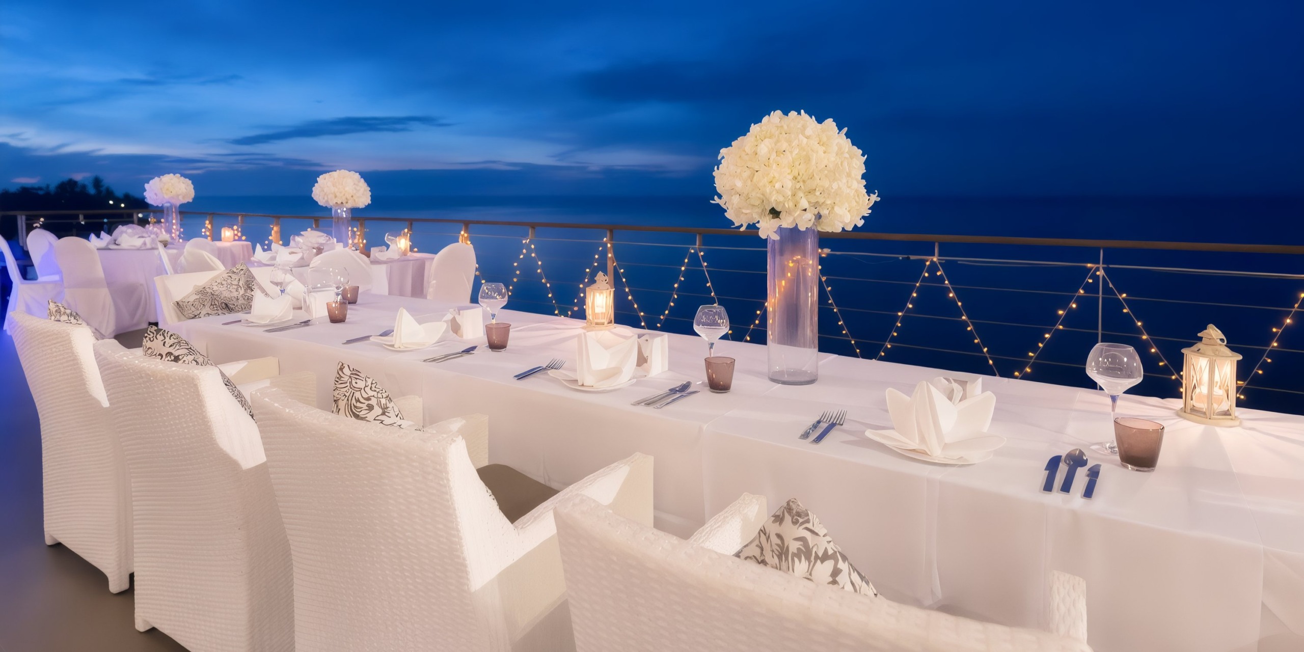 Experience an enchanting outdoor dining setup with the Princess Grand Jamaica, featuring white tablecloths, elegant floral centerpieces, and sparkling string lights, all overlooking the ocean at dusk.