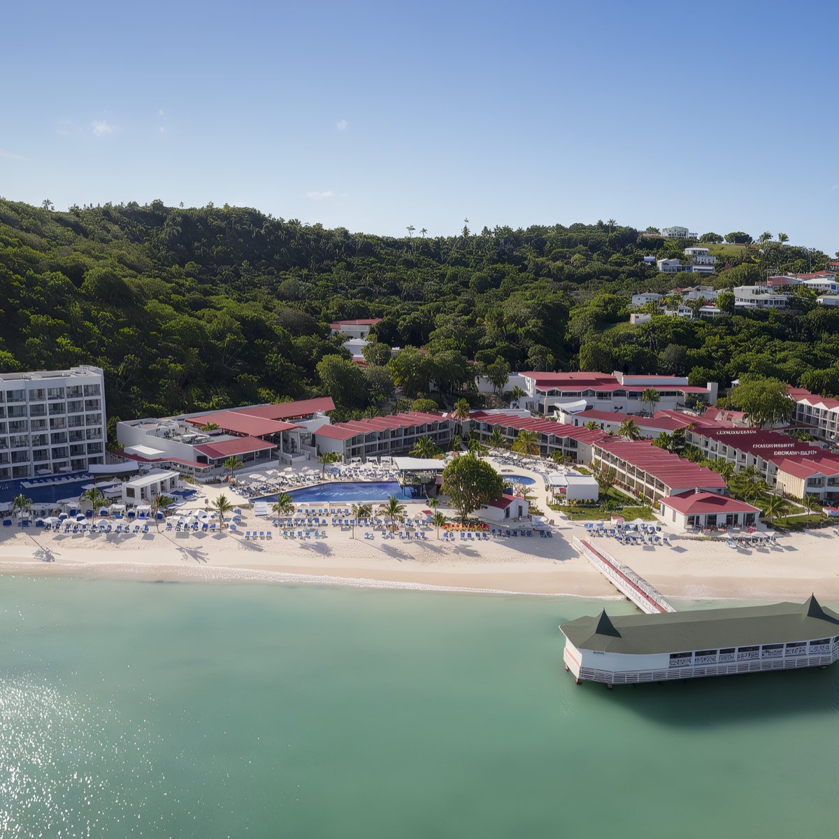 Aerial view of the Royalton Chic Antigua beachfront resort with red-roofed buildings, a white sandy beach, blue umbrellas, and a dock extending over calm green water, surrounded by dense green trees.