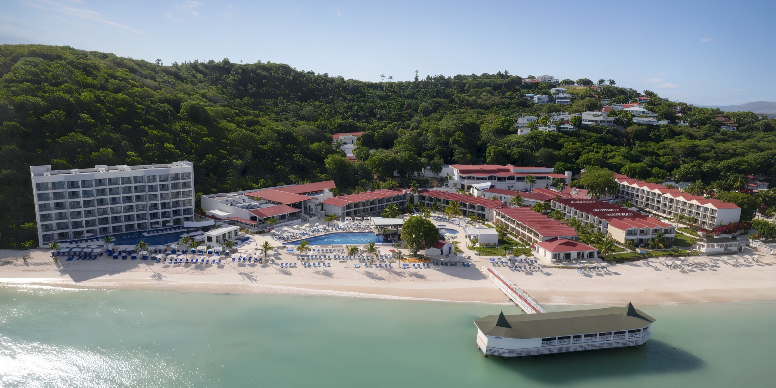Aerial view of the Royalton Chic Antigua beachfront resort, featuring multiple buildings, a swimming pool, and rows of blue beach chairs. Lush green hills stand tall in the background.