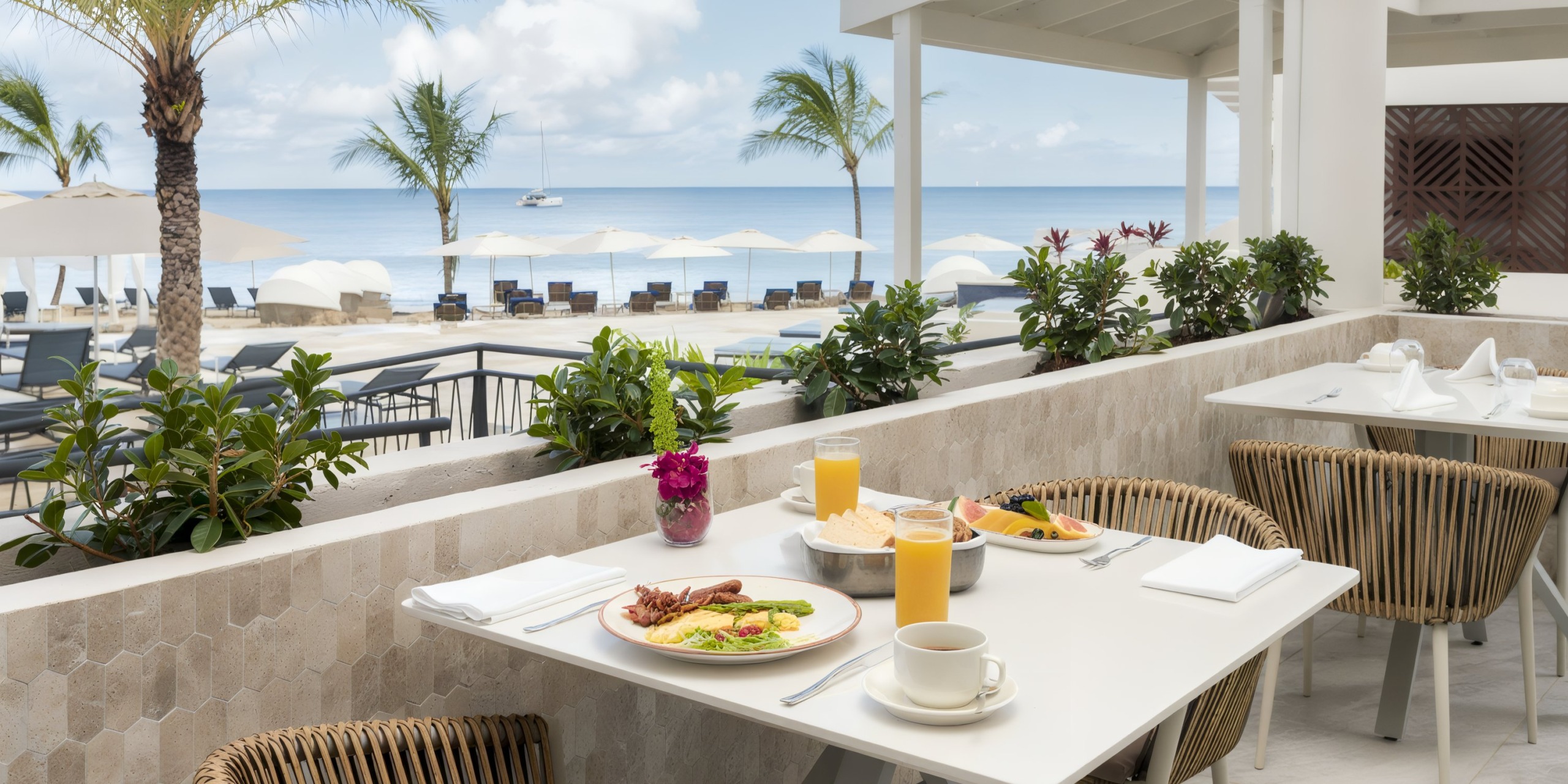Experience the Royalton Chic Antigua with a delightful outdoor dining setup featuring plates of food, refreshing juice, and smooth coffee on white tables overlooking a beach adorned with palm trees, lounge chairs, and a sailboat gliding across the ocean.