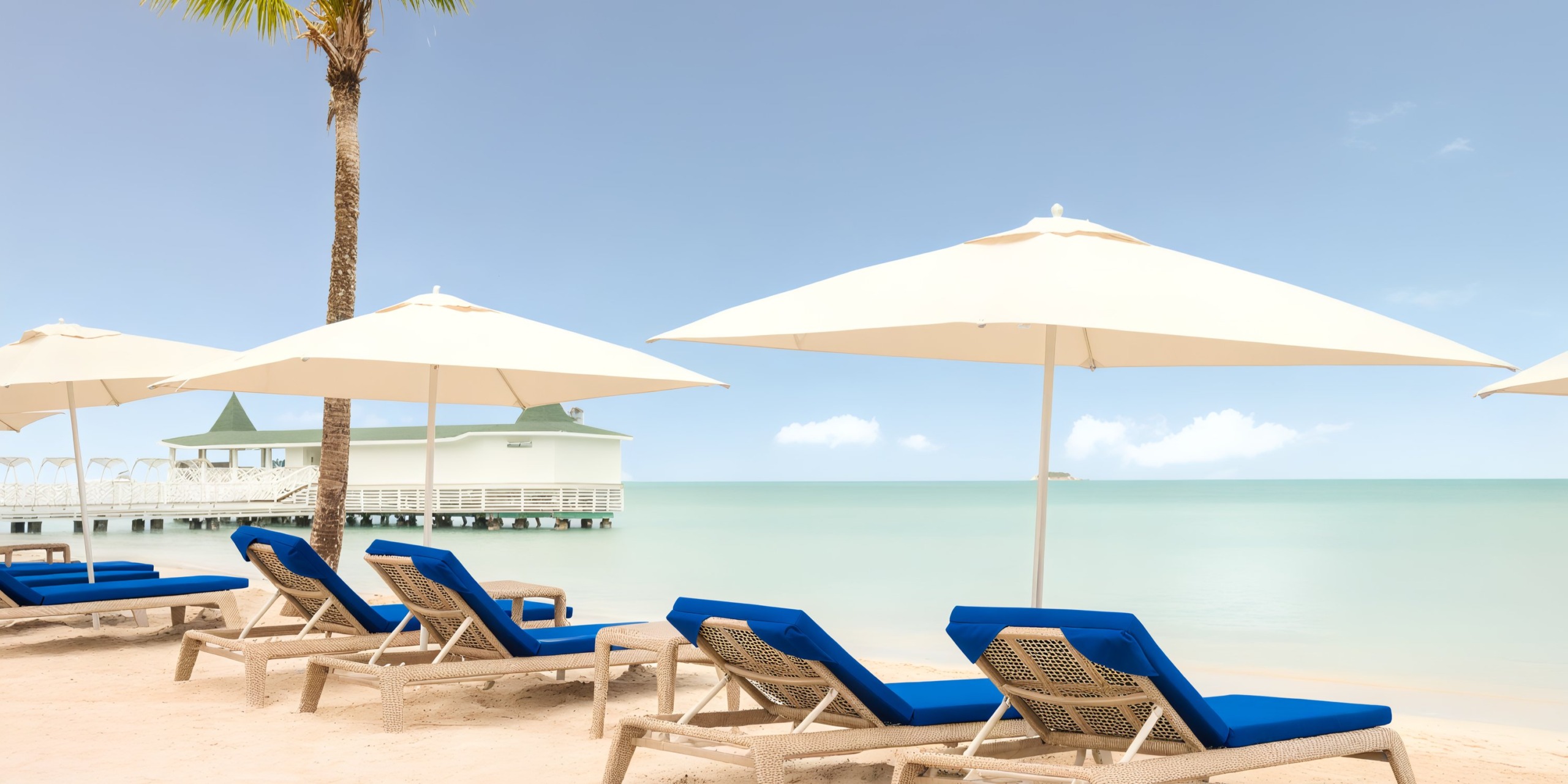 Scene at Royalton Chic Antigua: Lounge chairs with blue cushions are arranged under white umbrellas, facing a calm sea and a distant pier with a gazebo, complemented by a palm tree on the side.