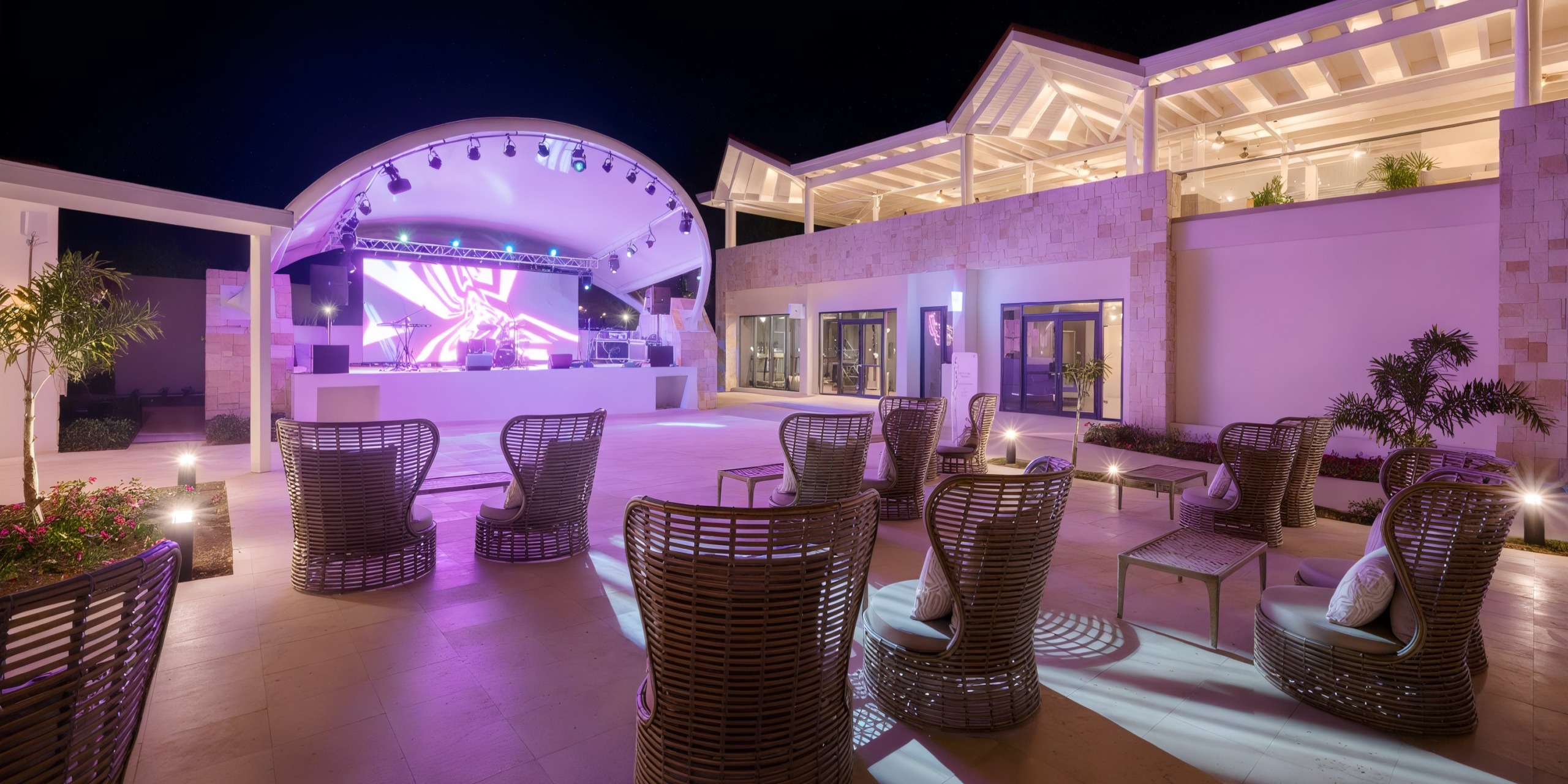 Outdoor lounge area at the Royalton Chic Antigua featuring wicker chairs facing a stage with a large screen display. The space is bathed in purple lighting and surrounded by modern architecture.