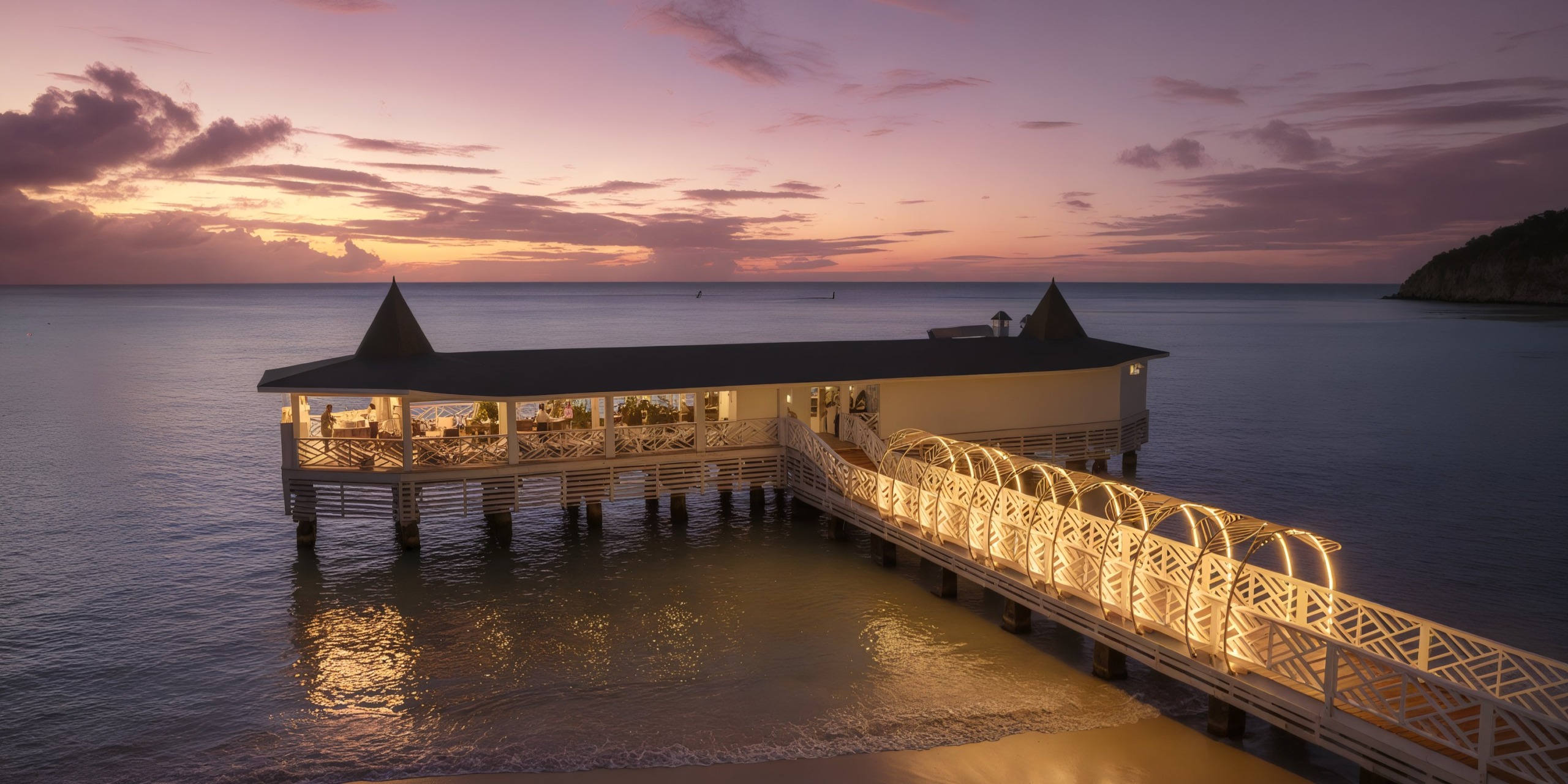 An illuminated walkway on the pier leads to Royalton Chic Antigua, a building situated over tranquil ocean waters beneath a sunset sky adorned with scattered clouds.