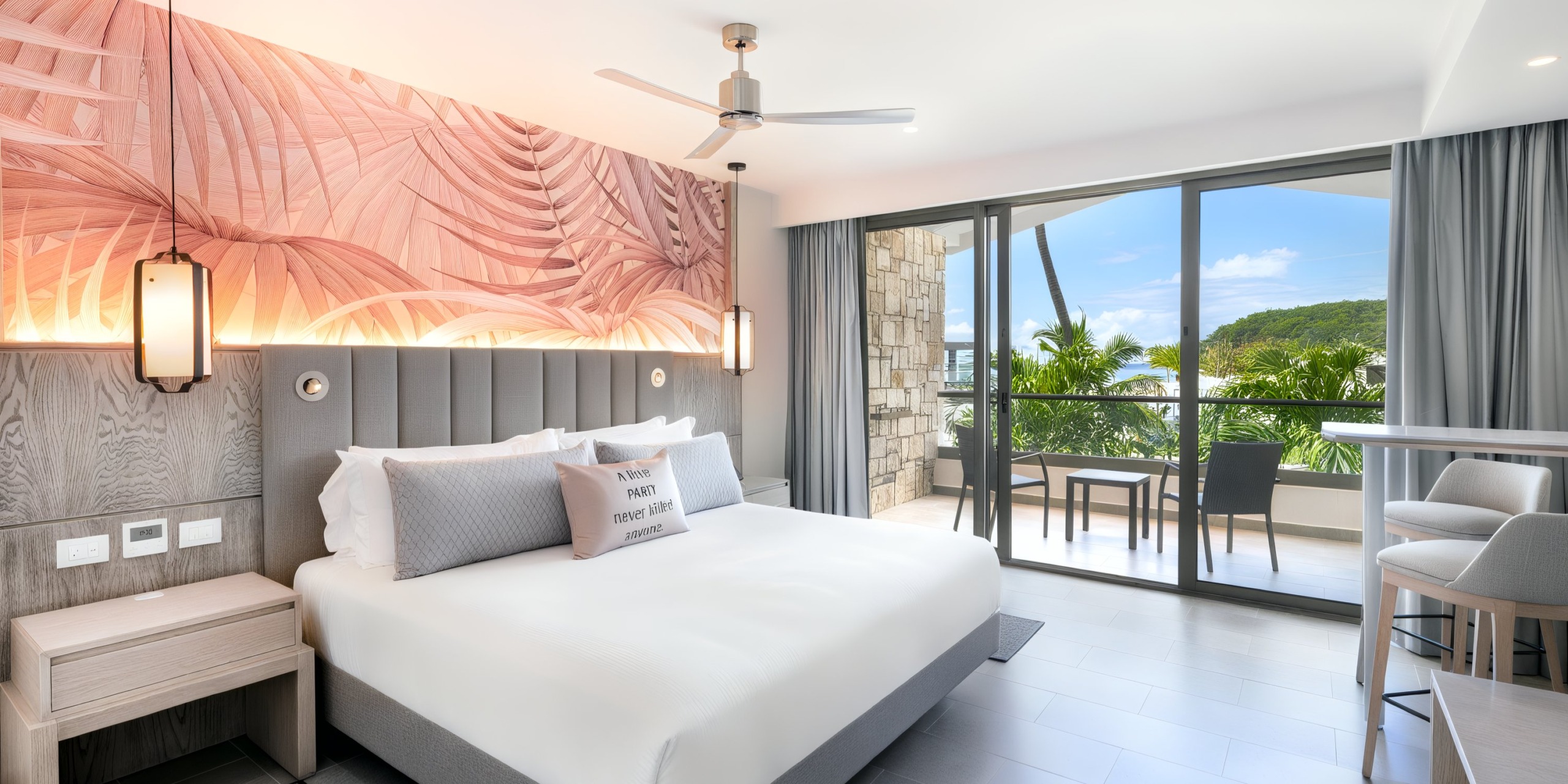 Experience a stay in the Royalton Chic Antigua, featuring a modern hotel room with a double bed, decorative wall art, and a balcony that offers stunning views of tropical scenery. Enjoy amenities such as a ceiling fan, bedside tables, and a comfortable seating area.